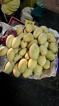 A pile of ripe, yellow mangoes placed on a paper-lined surface, with a handwritten price card displaying the number 50. In the background, there are plastic baskets and containers, highlighting a rustic market setting.