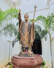 A bronze statue of a religious figure in traditional attire, holding a staff with a cross at the top. The figure is depicted with one hand raised in blessing. The statue is set against a white wall backdrop, surrounded by green palm plants, and positioned on a red granite pedestal.
