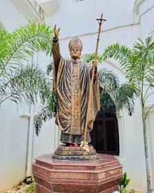 A bronze statue of a religious figure in traditional attire, holding a staff with a cross at the top. The figure is depicted with one hand raised in blessing. The statue is set against a white wall backdrop, surrounded by green palm plants, and positioned on a red granite pedestal.