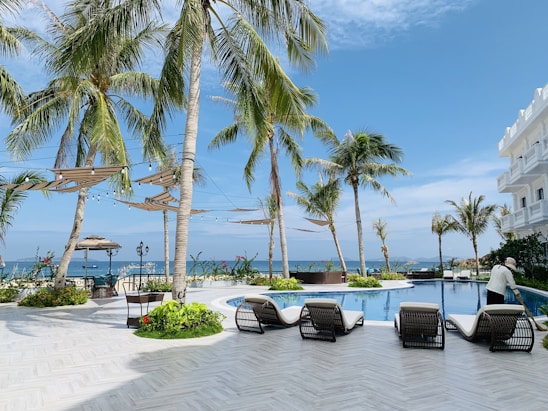 Palm trees surround a luxurious poolside area with lounge chairs on a sunny day. A person tends to the pool while decorative string lights hang above. The ocean can be seen in the background, and a white building stands to the right.