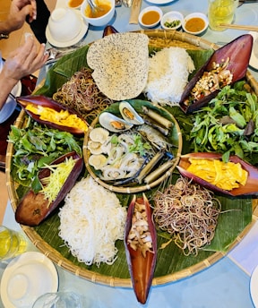 A large round bamboo tray is placed on a table, lined with banana leaves and filled with a variety of fresh ingredients, including different noodles, leafy greens, sliced fruits, shellfish, and rolls. Bowls of sauces and small dishes surround the tray. One person's hands are visible near the food, suggesting an interactive dining experience.