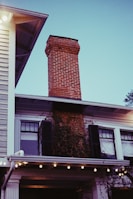 A brick chimney extends upwards from the roof of a two-story house. The house features white siding, black shutters, and ivy growing up the side. Strings of lights are hung under the eaves, creating a warm ambiance. The sky is clear with a bluish tint.