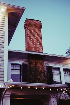 A brick chimney extends upwards from the roof of a two-story house. The house features white siding, black shutters, and ivy growing up the side. Strings of lights are hung under the eaves, creating a warm ambiance. The sky is clear with a bluish tint.