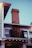 A brick chimney extends upwards from the roof of a two-story house. The house features white siding, black shutters, and ivy growing up the side. Strings of lights are hung under the eaves, creating a warm ambiance. The sky is clear with a bluish tint.