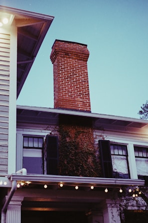 A brick chimney extends upwards from the roof of a two-story house. The house features white siding, black shutters, and ivy growing up the side. Strings of lights are hung under the eaves, creating a warm ambiance. The sky is clear with a bluish tint.