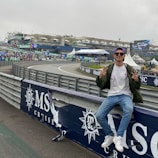 A person in casual attire sits on a barrier beside a racetrack, making peace signs with both hands. In the background, there is a stadium, a large crowd, promotional banners, and a screen displaying an advertisement. The scene has an overcast sky, and the setting appears to be a motorsport event.