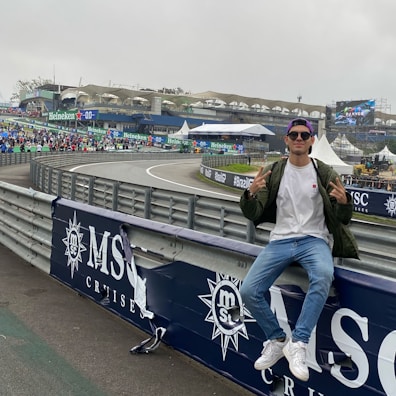 A person in casual attire sits on a barrier beside a racetrack, making peace signs with both hands. In the background, there is a stadium, a large crowd, promotional banners, and a screen displaying an advertisement. The scene has an overcast sky, and the setting appears to be a motorsport event.