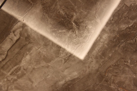 A close-up view of a textured marble or stone surface with a mix of dark and light brown shades. The surface features natural veining patterns and variations in color, creating an abstract, earthy look.