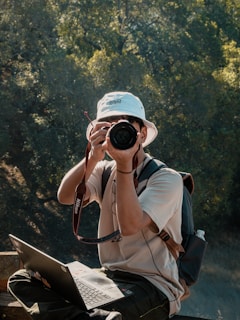 A person wearing a white bucket hat and a light-colored shirt is sitting outdoors with a laptop on their lap and holding a DSLR camera. The person has a backpack and is surrounded by lush green trees in a sunlit area.