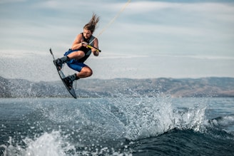 A person is performing an aerial maneuver on a wakeboard, holding onto a handle attached to a rope. There is a splash of water underneath as the wakeboarder is airborne, with a backdrop of calm water and distant hills under a cloudy sky.