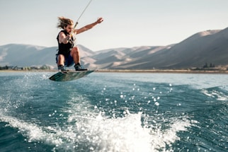 A person is performing an aerial maneuver on a wakeboard, with water splashing beneath. The scene is set on a wide, calm body of water surrounded by mountainous terrain under a clear sky.