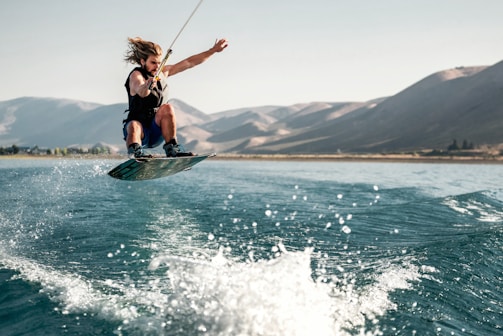 A person is performing an aerial maneuver on a wakeboard, with water splashing beneath. The scene is set on a wide, calm body of water surrounded by mountainous terrain under a clear sky.