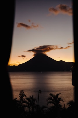 A silhouetted volcano stands prominently against a sunset sky, bordered by curtains. The sky transitions from warm orange tones to a deeper blue, and a few scattered clouds hover above the volcano's peak. The calm waters of a lake reflect the sky's colors, and palm trees frame the lower foreground.