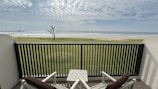Two comfortable chairs with brown cushions face a small table on an outdoor balcony, overlooking a serene beachfront landscape. A well-manicured green lawn stretches toward a sandy beach, bordered by a few small palm trees. In the distance, gentle waves ripple under a partly cloudy sky, creating a peaceful and relaxing atmosphere.