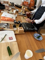 Several individuals participate in a cooking class, preparing ingredients on wooden tables. Cutting boards, knives, bottles of condiments, and recipe sheets are visible. Some participants wear aprons, and a variety of kitchen utensils are scattered around.