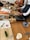 Several individuals participate in a cooking class, preparing ingredients on wooden tables. Cutting boards, knives, bottles of condiments, and recipe sheets are visible. Some participants wear aprons, and a variety of kitchen utensils are scattered around.