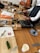 Several individuals participate in a cooking class, preparing ingredients on wooden tables. Cutting boards, knives, bottles of condiments, and recipe sheets are visible. Some participants wear aprons, and a variety of kitchen utensils are scattered around.