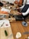 Several individuals participate in a cooking class, preparing ingredients on wooden tables. Cutting boards, knives, bottles of condiments, and recipe sheets are visible. Some participants wear aprons, and a variety of kitchen utensils are scattered around.