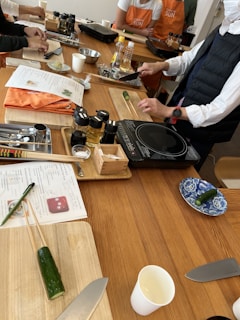 Several individuals participate in a cooking class, preparing ingredients on wooden tables. Cutting boards, knives, bottles of condiments, and recipe sheets are visible. Some participants wear aprons, and a variety of kitchen utensils are scattered around.