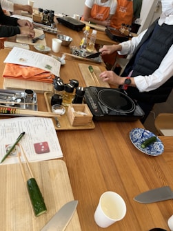 Several individuals participate in a cooking class, preparing ingredients on wooden tables. Cutting boards, knives, bottles of condiments, and recipe sheets are visible. Some participants wear aprons, and a variety of kitchen utensils are scattered around.