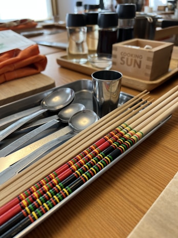 A set of colorful chopsticks, each decorated with intricate patterns in red, yellow, green, and black, is neatly arranged in a tray on a wooden table. The table also features metal spoons, measuring spoons, a metallic cup, and various containers with condiments. A box with the label 'Cooking Sun' is present in the background.
