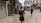 A person walks through a bustling market street lined with various shops. The shops display signs for currency exchange and an ATM. There are several other pedestrians further along the cobblestone street, which is bordered by historic buildings.