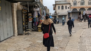 A person walks through a bustling market street lined with various shops. The shops display signs for currency exchange and an ATM. There are several other pedestrians further along the cobblestone street, which is bordered by historic buildings.