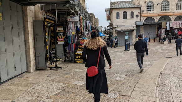 A person walks through a bustling market street lined with various shops. The shops display signs for currency exchange and an ATM. There are several other pedestrians further along the cobblestone street, which is bordered by historic buildings.