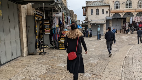 A person walks through a bustling market street lined with various shops. The shops display signs for currency exchange and an ATM. There are several other pedestrians further along the cobblestone street, which is bordered by historic buildings.