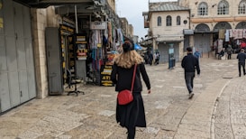 A person walks through a bustling market street lined with various shops. The shops display signs for currency exchange and an ATM. There are several other pedestrians further along the cobblestone street, which is bordered by historic buildings.