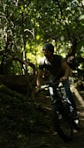A person wearing a helmet is riding a fat-tire bike through a dense, leafy forest. The sunlight filters through the trees, casting dappled light on the ground and creating a serene, adventurous atmosphere.