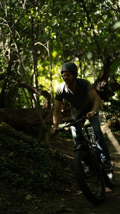 A person wearing a helmet is riding a fat-tire bike through a dense, leafy forest. The sunlight filters through the trees, casting dappled light on the ground and creating a serene, adventurous atmosphere.