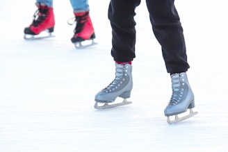 Two individuals ice skating, with a focus on their legs and skates. One person is wearing gray skates and black pants, while the other sports red skates with jeans. The scene captures movement and a sense of fun on the ice.