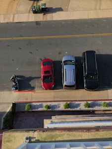 An overhead view of a street scene featuring three parked vehicles: a red car, a silver van, and a dark SUV. Adjacent to the vehicles, a person is either mounting or dismounting a motorcycle. The street is bordered by a sidewalk and a row of small, evenly spaced shrubs. Trash bins are positioned at the top left corner on a concrete section.