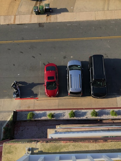 An overhead view of a street scene featuring three parked vehicles: a red car, a silver van, and a dark SUV. Adjacent to the vehicles, a person is either mounting or dismounting a motorcycle. The street is bordered by a sidewalk and a row of small, evenly spaced shrubs. Trash bins are positioned at the top left corner on a concrete section.