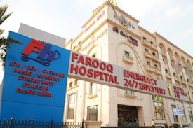 A hospital building with an entrance sign indicating various departments such as ICU, CCU, Cath Lab, Paediatrics, Nursery, Stroke Unit, Dialysis, and Blood Bank. The building has a beige facade with multiple stories and several windows. There are various signs with red and blue lettering highlighting emergency services and 24/7 inpatient care.