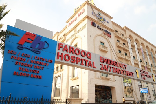 A hospital building with an entrance sign indicating various departments such as ICU, CCU, Cath Lab, Paediatrics, Nursery, Stroke Unit, Dialysis, and Blood Bank. The building has a beige facade with multiple stories and several windows. There are various signs with red and blue lettering highlighting emergency services and 24/7 inpatient care.