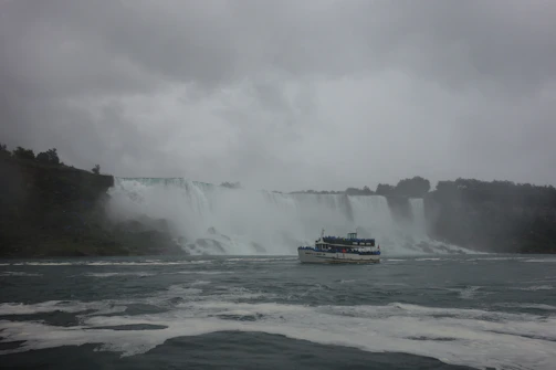 A large waterfall cascades over a wide cliff with lush greenery on either side. A tour boat is sailing in the choppy waters below, filled with passengers in blue raincoats. The sky above is overcast with thick grey clouds, creating a misty atmosphere.