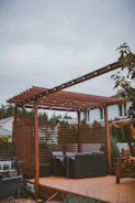 A wooden pergola with string lights is situated on a patio. Beneath the pergola, there are cushioned outdoor chairs arranged around a small table. The area is surrounded by a variety of plants, including some trees and flowering bushes. In the background, there are residential houses with green and brown roofs visible.