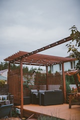 A wooden pergola with string lights is situated on a patio. Beneath the pergola, there are cushioned outdoor chairs arranged around a small table. The area is surrounded by a variety of plants, including some trees and flowering bushes. In the background, there are residential houses with green and brown roofs visible.