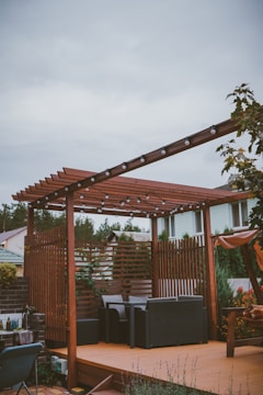 A wooden pergola with string lights is situated on a patio. Beneath the pergola, there are cushioned outdoor chairs arranged around a small table. The area is surrounded by a variety of plants, including some trees and flowering bushes. In the background, there are residential houses with green and brown roofs visible.