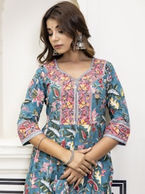 A woman wearing a blue and pink floral-patterned traditional dress stands against a light-colored wall. She has long brown hair, wearing statement silver jhumka earrings, and has her hands gently crossed. The dress features intricate designs with vibrant flowers, leaves, and geometric patterns.