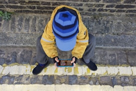 A person sitting on a cobblestone pavement with a stone wall in the background is holding a smartphone to play a game. The individual is wearing a yellow jacket, gray pants, dark shoes, and a blue hat with horizontal stripes.
