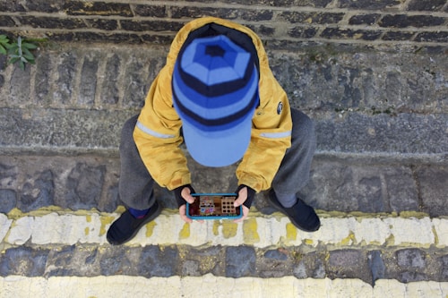 A person sitting on a cobblestone pavement with a stone wall in the background is holding a smartphone to play a game. The individual is wearing a yellow jacket, gray pants, dark shoes, and a blue hat with horizontal stripes.