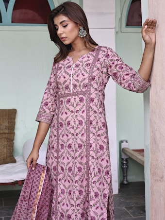 A woman stands in a relaxed pose, wearing a pink floral-patterned traditional dress. Her long, dark hair is styled down and she is adorned with large, ornate earrings. The backdrop features soft, pastel-colored walls and a cushioned bench. The overall setting exudes a calm and elegant atmosphere.