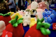 A collection of colorful plush toys displayed on a surface. Various stuffed animals, including a white unicorn with a horn and blue elephants, are surrounded by plush strawberries and other toys. Several people are visible in the background, out of focus.