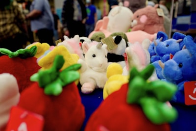 A collection of colorful plush toys displayed on a surface. Various stuffed animals, including a white unicorn with a horn and blue elephants, are surrounded by plush strawberries and other toys. Several people are visible in the background, out of focus.