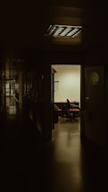 A dimly lit hallway leading to an illuminated office with an open door. Inside the office, there is a desk with a chair, and papers scattered on the desk. The polished floor reflects some light, and shadows dominate the corridor.