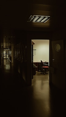 A dimly lit hallway leading to an illuminated office with an open door. Inside the office, there is a desk with a chair, and papers scattered on the desk. The polished floor reflects some light, and shadows dominate the corridor.