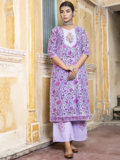A woman in traditional attire stands against a richly detailed, textured wall. She wears a purple floral patterned kurta with matching palazzo pants, accessorized with a chunky metallic necklace and earrings. Her hair is neatly tied back, and she holds a smartphone in her hand.